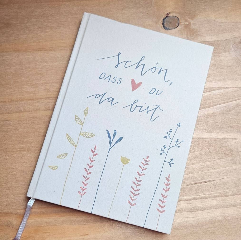 Gästebuch des Ferienhauses Pathe mit dem Cover-Schriftzug „Schön, dass du da bist“ und gezeichneten Gräsern und Blumen auf einem Holztisch